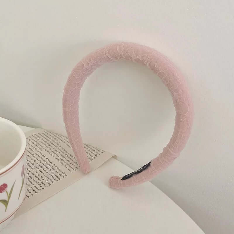A single pale pink sheer ruffle-textured padded headband resting on an open book beside a floral mug.