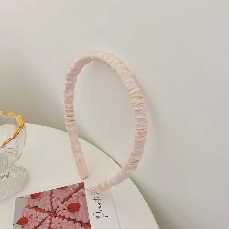 A single cream ivory ruched fabric headband styled upright near a glass dish and small photo card.