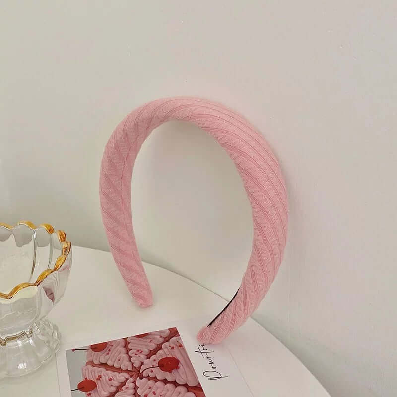 A single pink cable-knit textured padded headband styled upright near a glass dish and photo card.