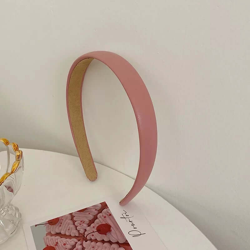 A single solid rose pink glossy padded headband styled upright near a glass dish and small photo card.
