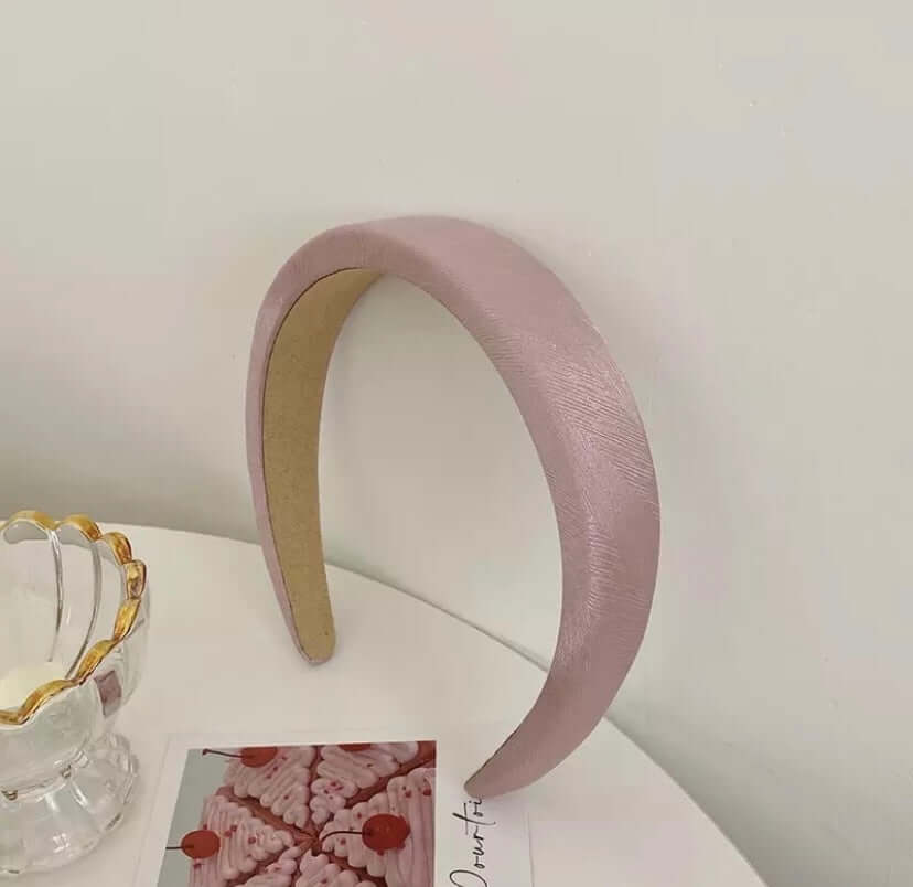 A single lilac mauve shimmer satin padded headband styled upright near a glass dish and photo card.