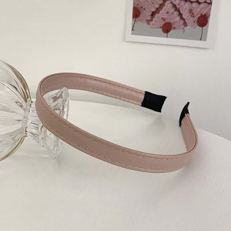 A single tan nude patent leather stitched headband styled near a glass ornament and small photo card.