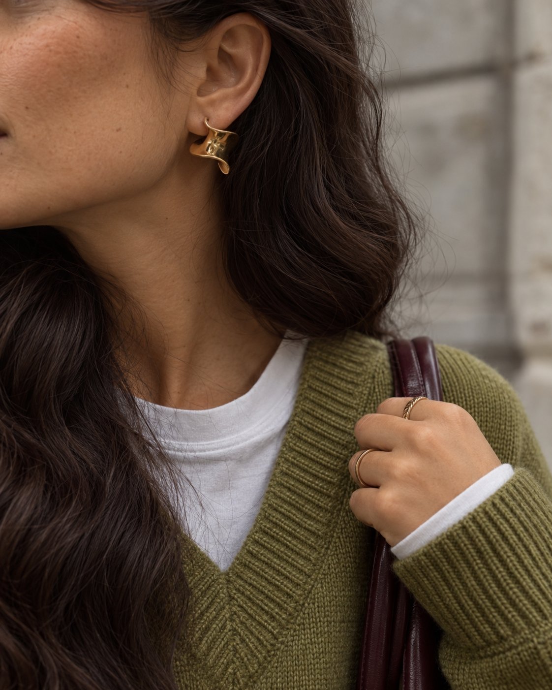Woman in an olive V-neck knit and white tee wearing gold Ribbon EARRINGS, holding a burgundy bag strap — Folie à Trois