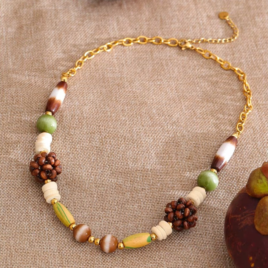 The Rio NECKLACE laid on woven fabric beside a carved wooden figure, its jade and wood cluster beads shown in close detail.