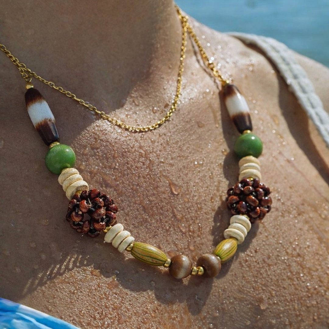 The Rio NECKLACE worn close-up on sun-damp skin, layered with a thin gold chain against a bright ocean backdrop.