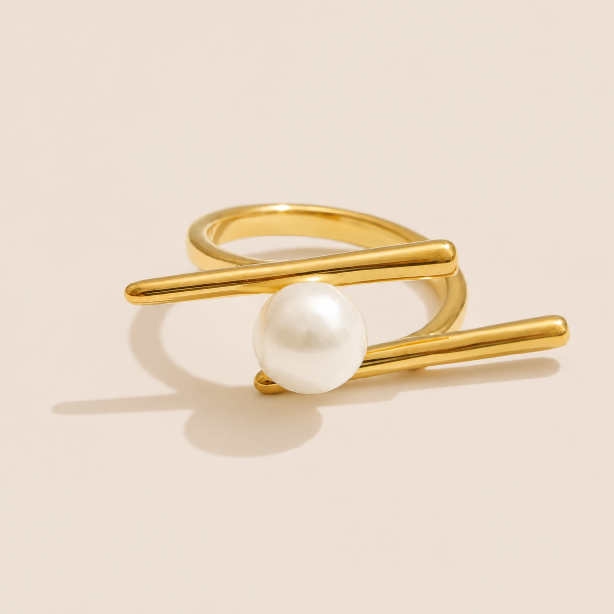 Gold double-band ring with two crossing bars cradling a single round pearl, shown on a cream backdrop — Folie à Trois Riviera Club RING.