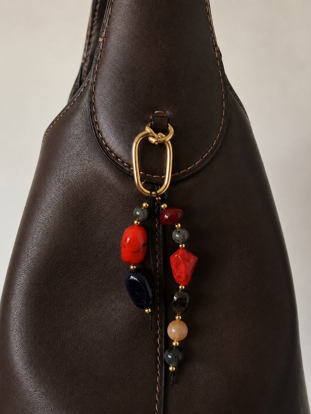 Folie à Trois Rocca BAG CHARM in red, black and grey natural stone beads, clipped to a dark brown leather bucket bag.