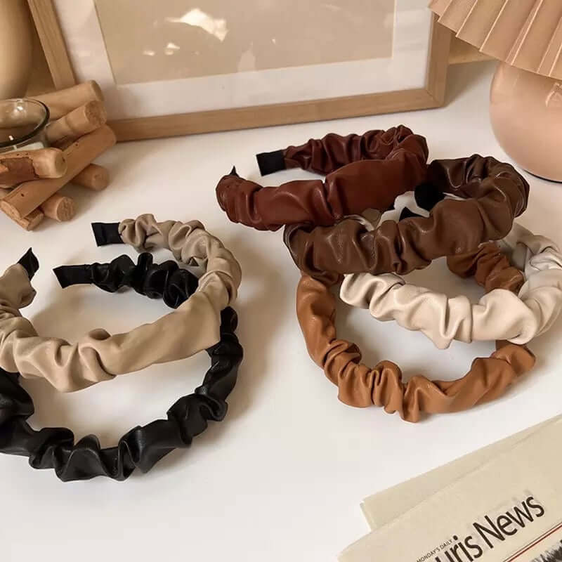 Five Romy HAIRBAND ruched faux leather headbands in beige, black, chestnut and cream, grouped on a white surface near a frame and lamp.