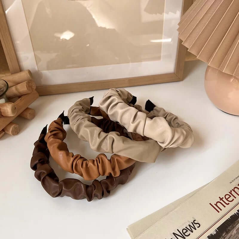 Three Romy HAIRBAND ruched faux leather headbands in cream, camel and dark brown, arranged together near a picture frame and lamp.