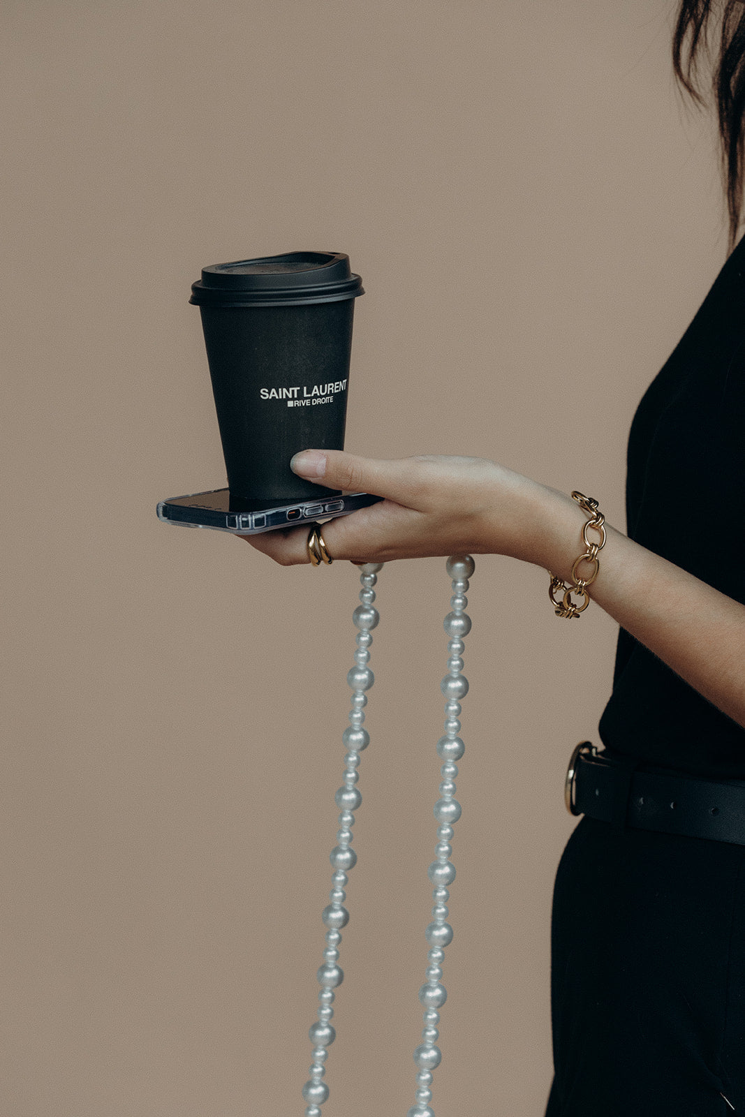 Close-up of a model's hand holding a phone with the Folie à Trois Sabine Pearl phone chain draped from it and a coffee cup balanced on top.