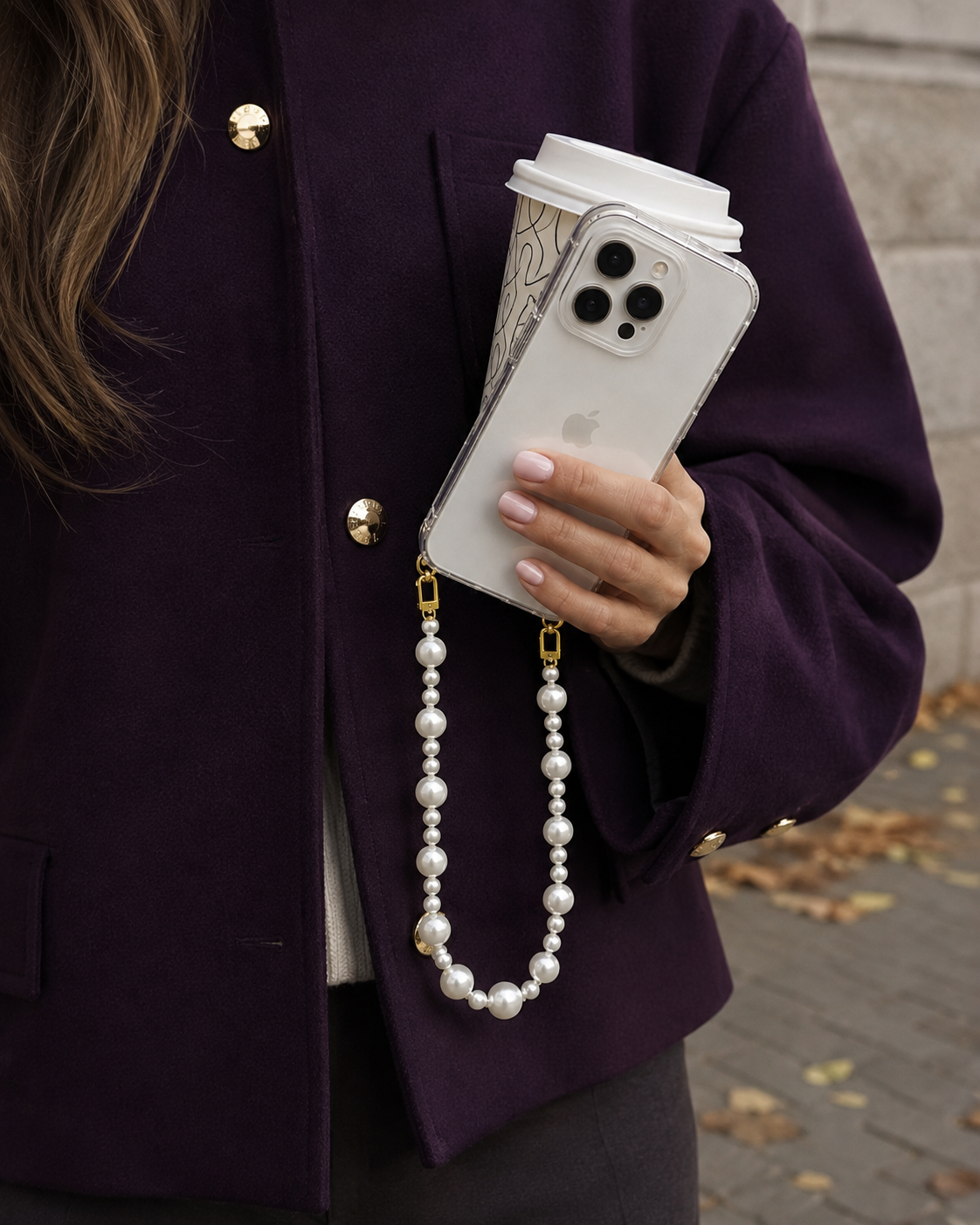 Model in a purple coat holding a coffee cup and phone with the Folie à Trois Sabine Pearl wholesale phone chain attached to the case.