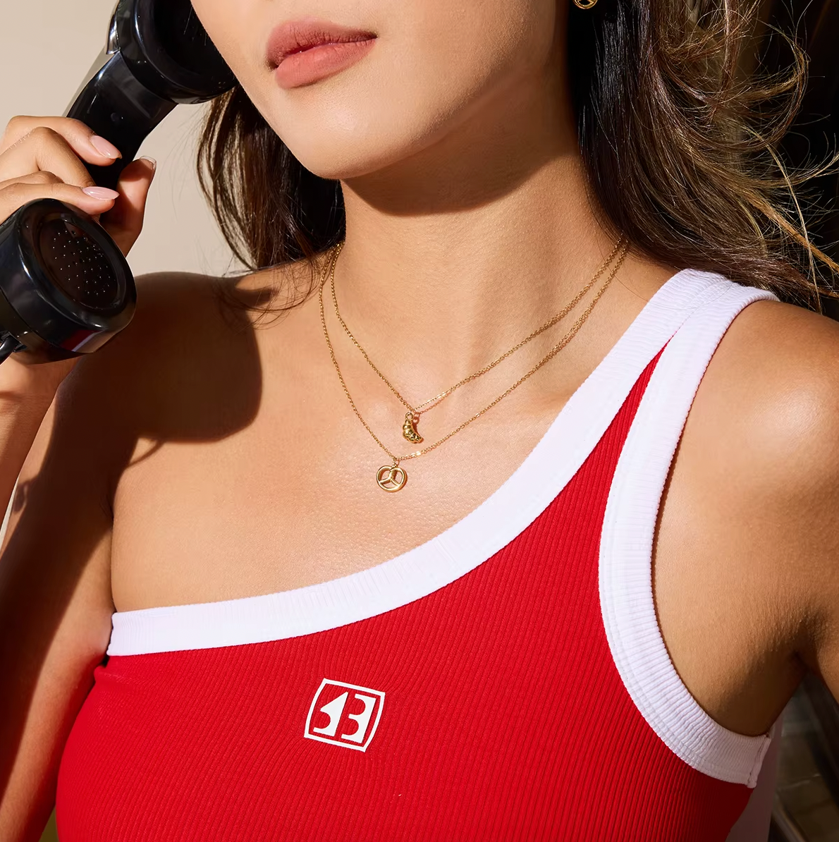 The Salty Sweet NECKLACE worn layered with a croissant charm necklace on a one-shoulder red top, phone held to her ear.