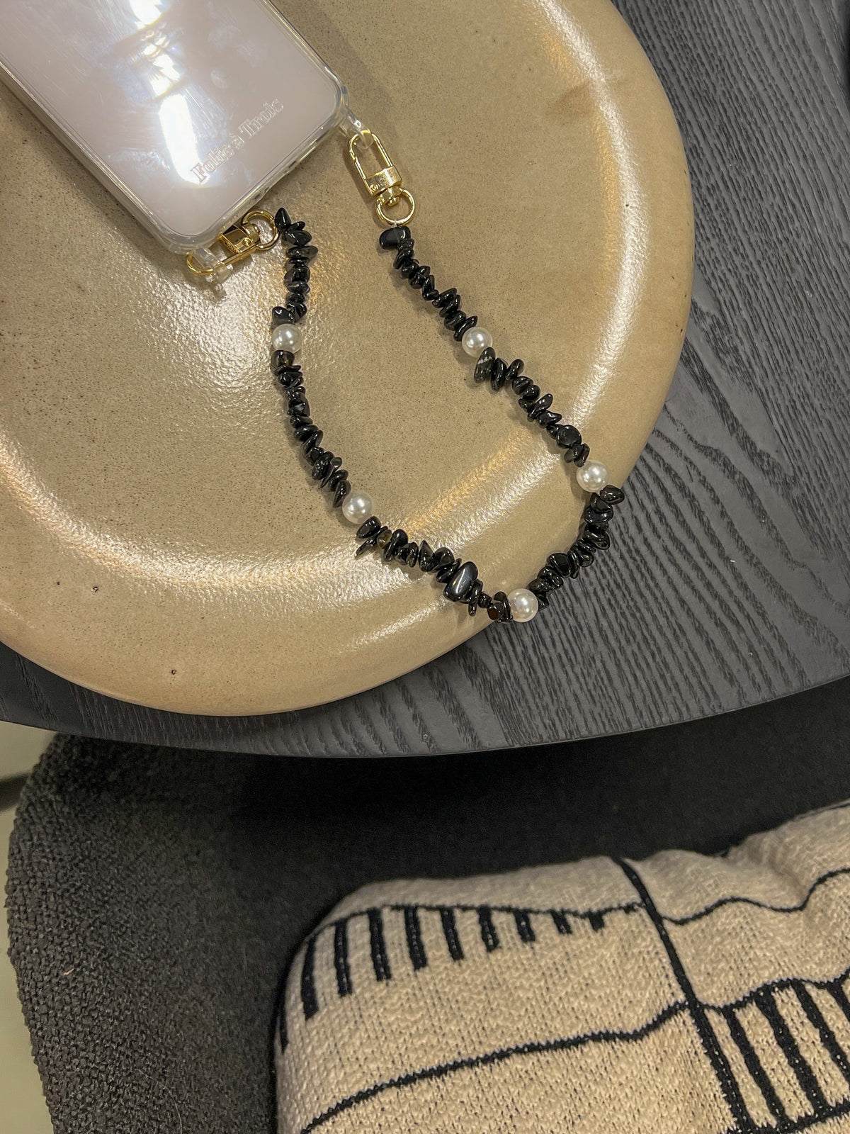 Black beaded Shell Whisper SHORT PHONE STRAP attached to a clear phone case, styled on a tan plate with dark textile background.