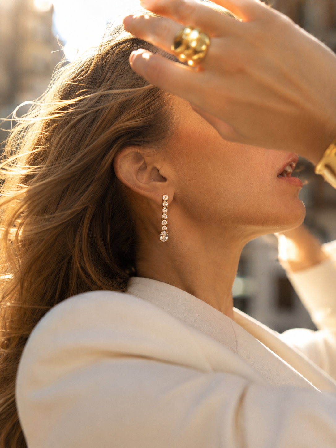 Model shields her eyes on a city street, wearing the gold Shimmer EARRINGS graduated zirconia drop with stacked gold rings.
