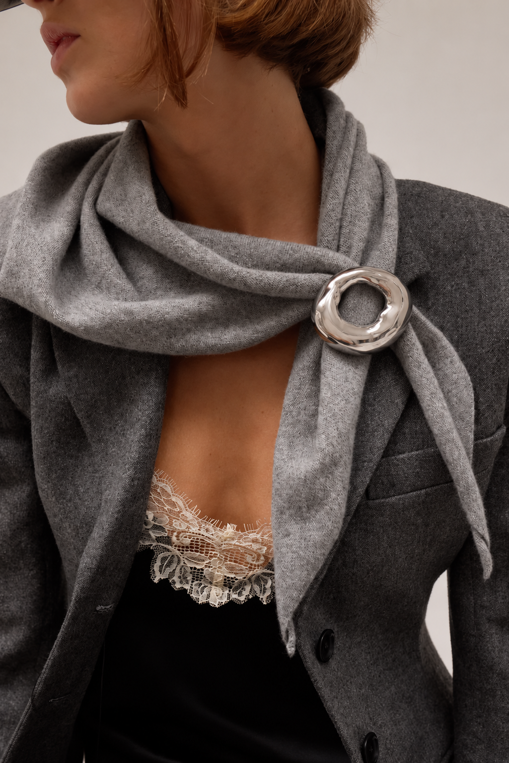 Sculptural silver-tone ring brooch pinning a grey wool scarf over a tailored grey blazer and lace top — Folie à Trois