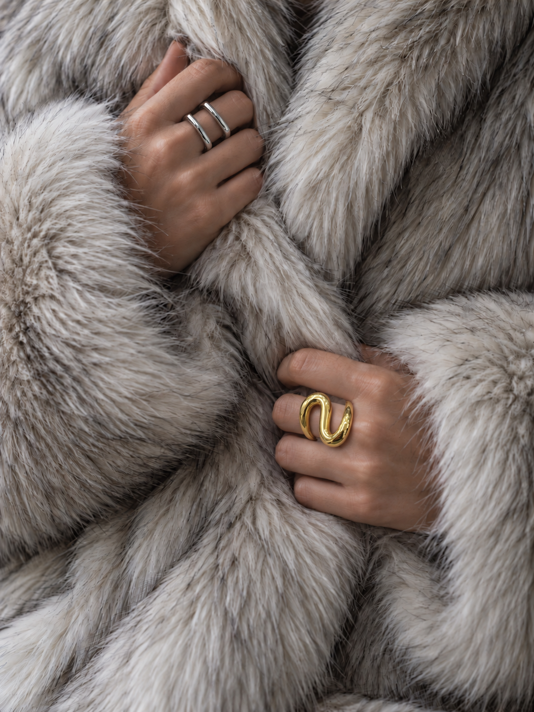 Hands clutching a grey fur coat, one wearing the gold S-curve Sinuosa RING, the other silver-tone rings — Folie à Trois