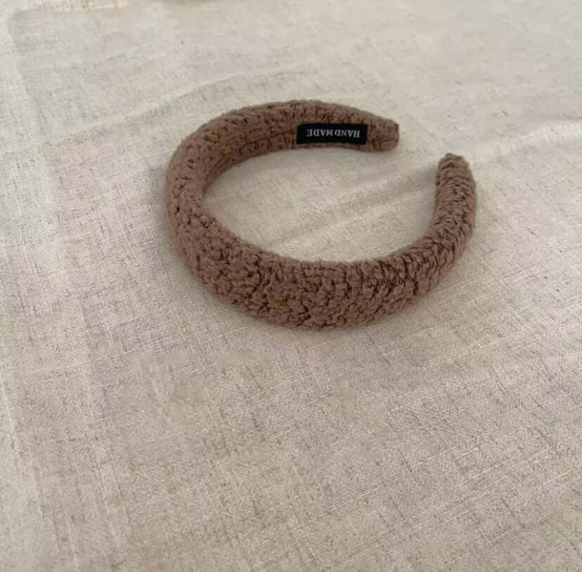 A single taupe brown boucle padded Skylar HAIRBAND with a handmade label tag, laid on linen fabric.