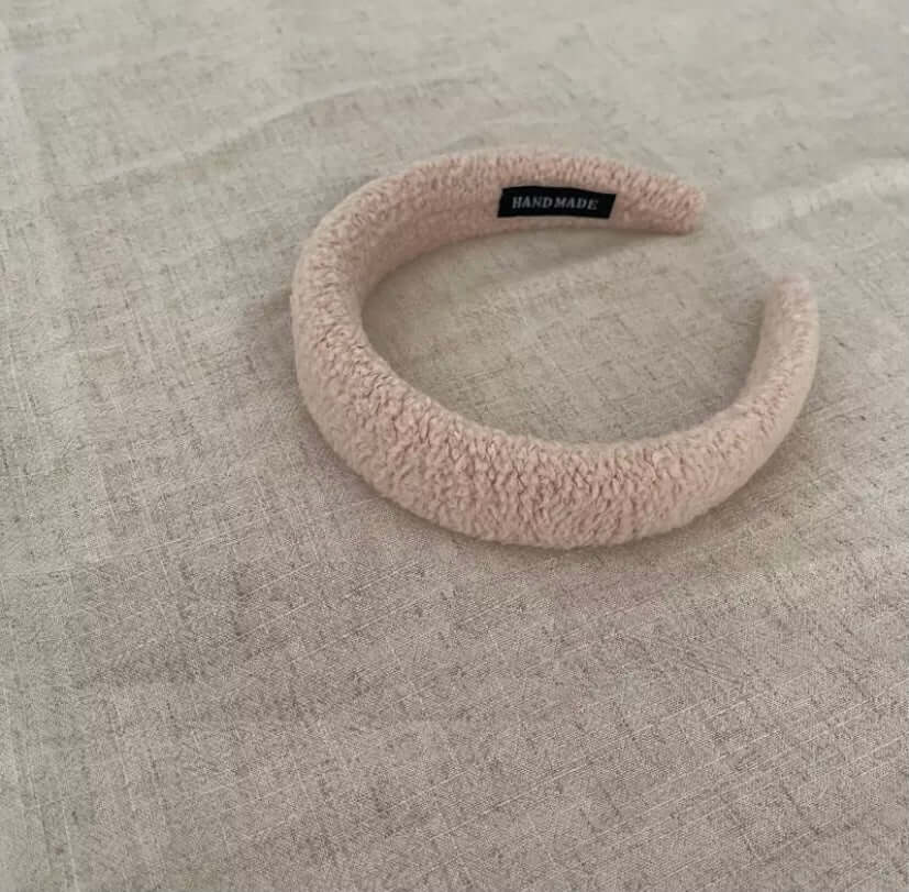 A single blush pink boucle padded Skylar HAIRBAND with a handmade label tag, laid on linen fabric.