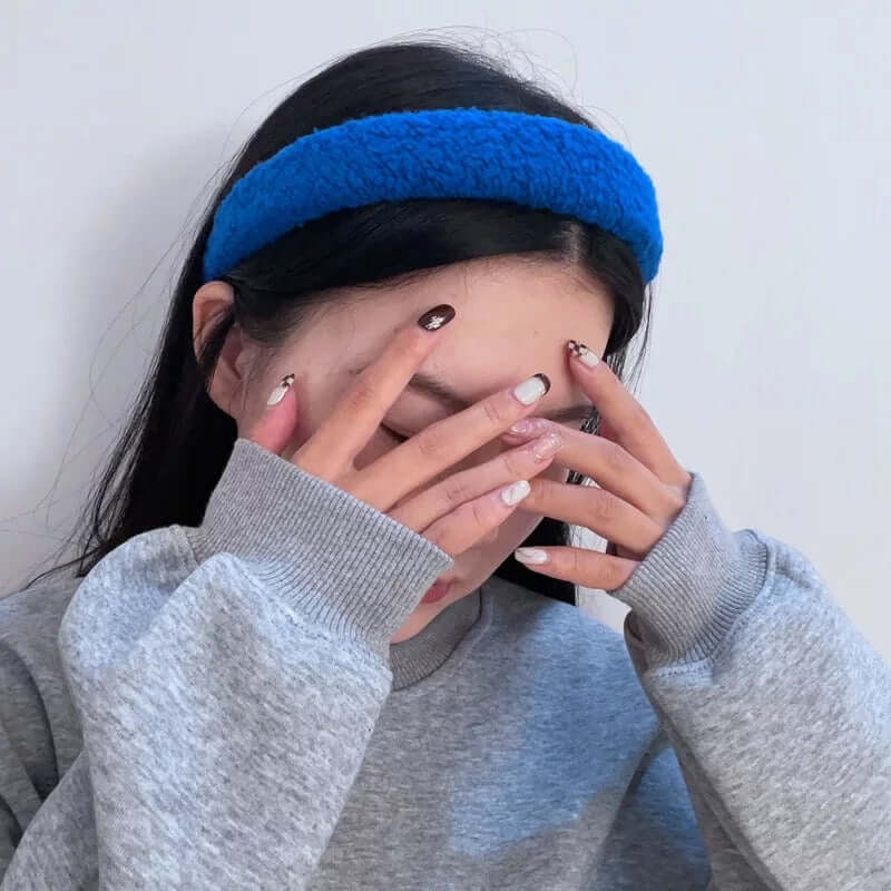 Close-up of a young woman wearing a cobalt blue boucle padded Skylar HAIRBAND, hands covering her face.