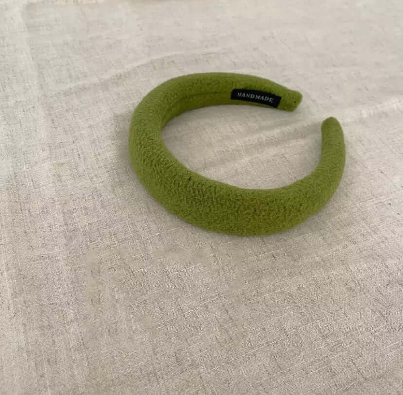 A single olive green boucle padded Skylar HAIRBAND with a handmade label tag, laid on linen fabric.