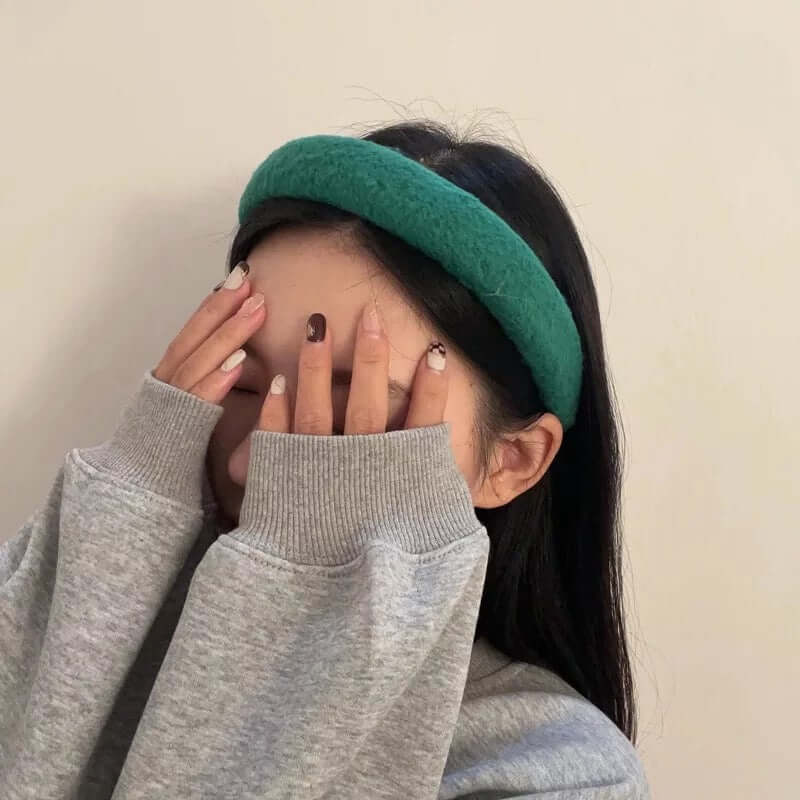 A young woman wearing a teal green boucle padded Skylar HAIRBAND, with her hand raised covering her face.