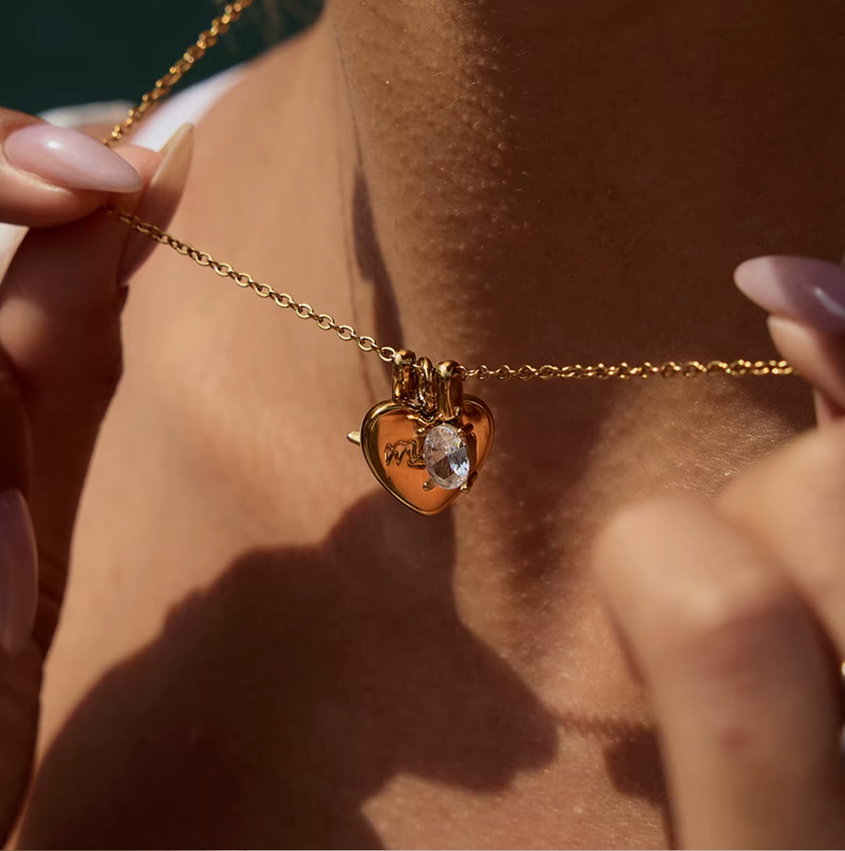 The Soft Power NECKLACE held between two fingers in extreme macro close-up, showing the engraved mom heart and crystal charm.