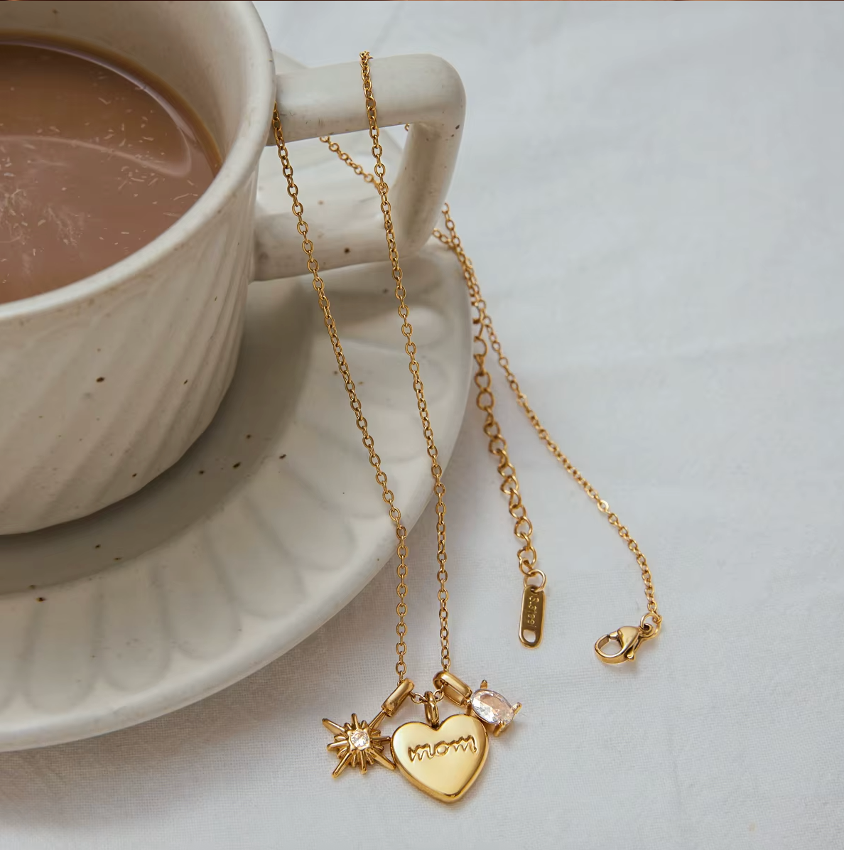The Soft Power NECKLACE draped over a teacup handle, its charms and gold clasp visible beside a cup of coffee.