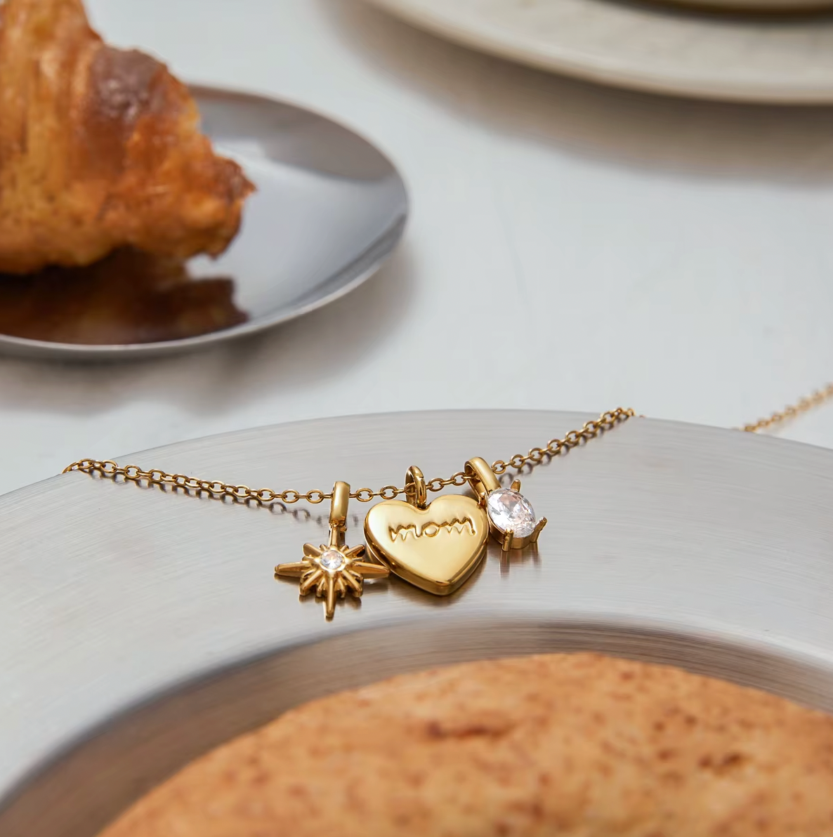 The Soft Power NECKLACE laid on a plate beside a croissant, its star, heart, and crystal charms shown in close detail.