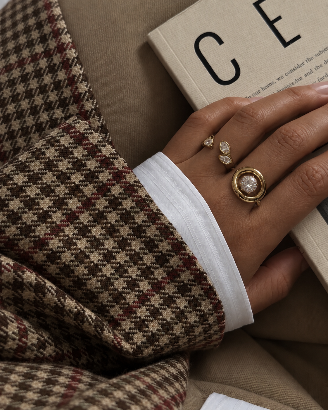 A hand in a houndstooth blazer sleeve rests on a book, wearing the Folie à Trois Solene RING stacked with two smaller pavé rings.