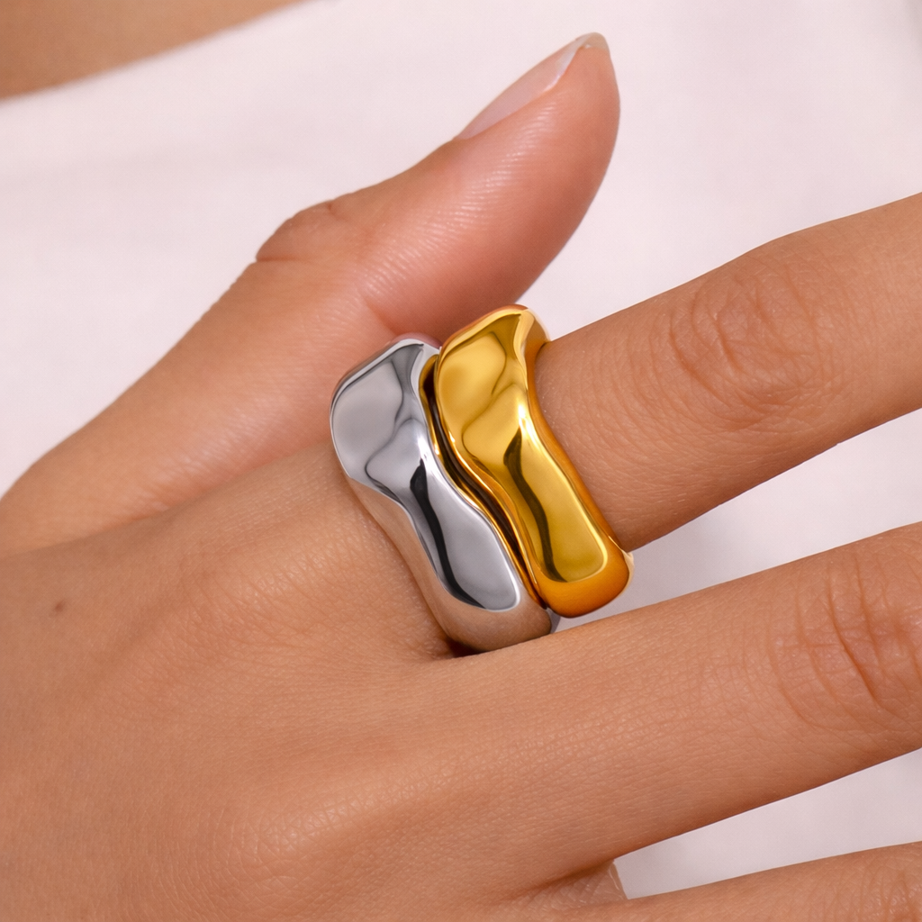 Close-up of a hand wearing two stacked wavy rings, one silver and one gold, each with an asymmetric dipped band shape.