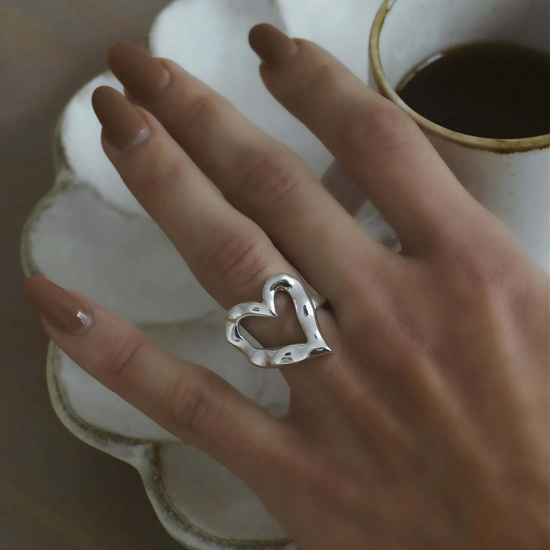 Close-up of a hand wearing the open silver heart-shaped Street Side RING, resting near a coffee cup on a scalloped plate.