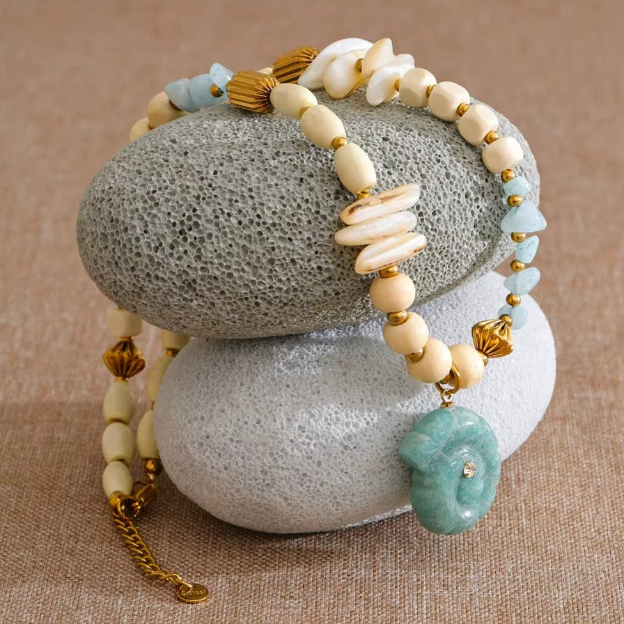 The Tahiti NECKLACE draped over two textured stones, showing its wood beads, shell chips, and carved amazonite pendant in detail.