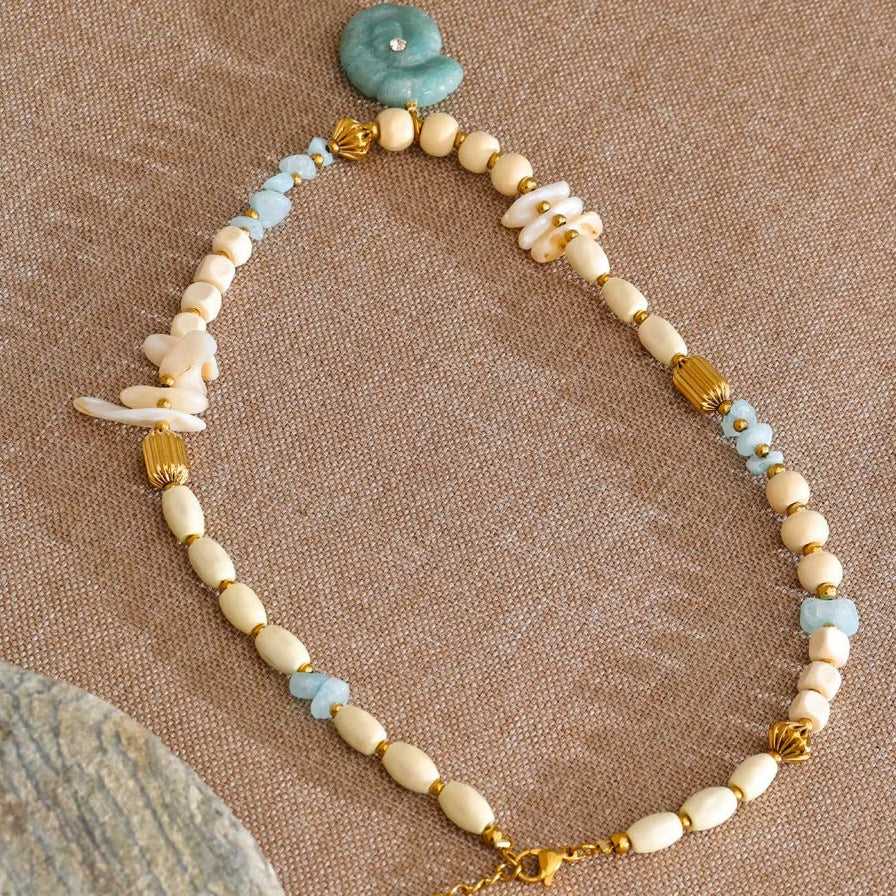 The Tahiti NECKLACE laid flat on a woven surface beside a stone, its shell chips and carved amazonite pendant on full display.