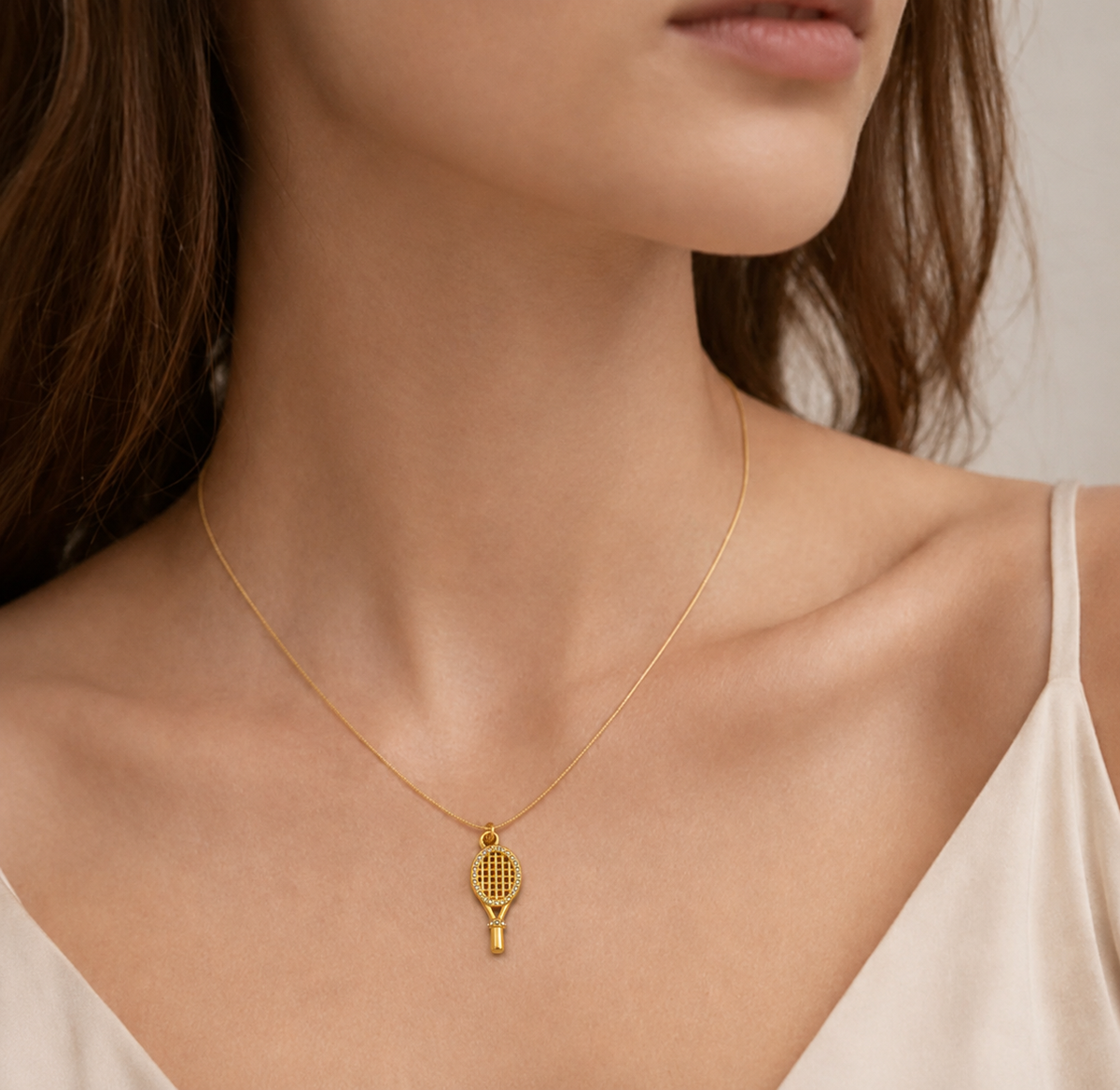 A close-up of a model wearing a delicate gold tennis racket pendant necklace on a fine chain, paired with a light beige camisole top.