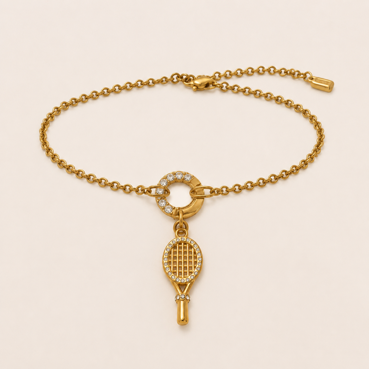 A gold chain bracelet featuring a tennis racket charm encrusted with small clear stones, displayed on a light beige background.