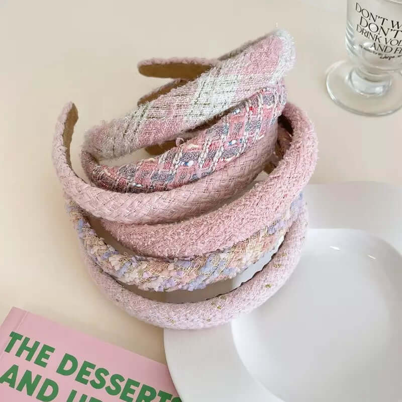 Six pink-toned tweed headbands in different patterns stacked together on a plate near a book.
