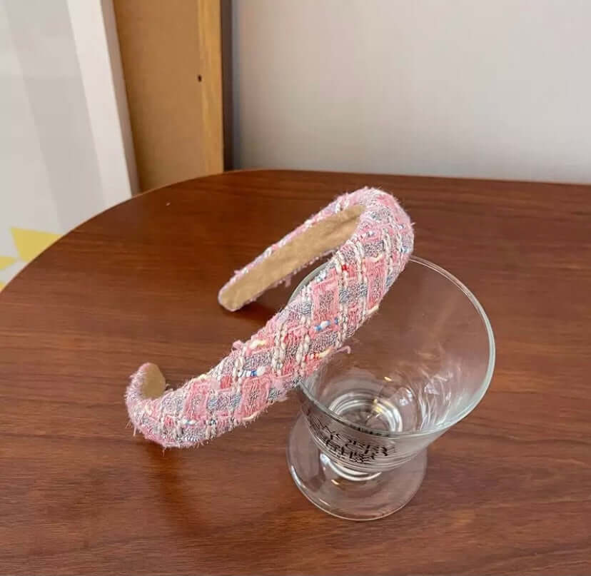 A single pink, grey and white tweed headband with small bead accents resting on a wood table beside a glass.