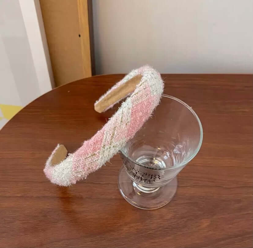 A single pink and white sparkly diamond-weave tweed headband resting on a wood table beside a glass.
