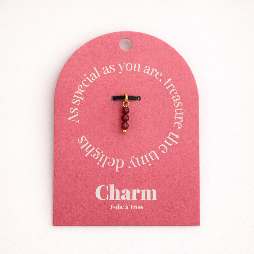 The Three Beads CHARM displayed on a coral pink Charm gift tag, three faceted deep red-toned glass beads hanging from a black cord.
