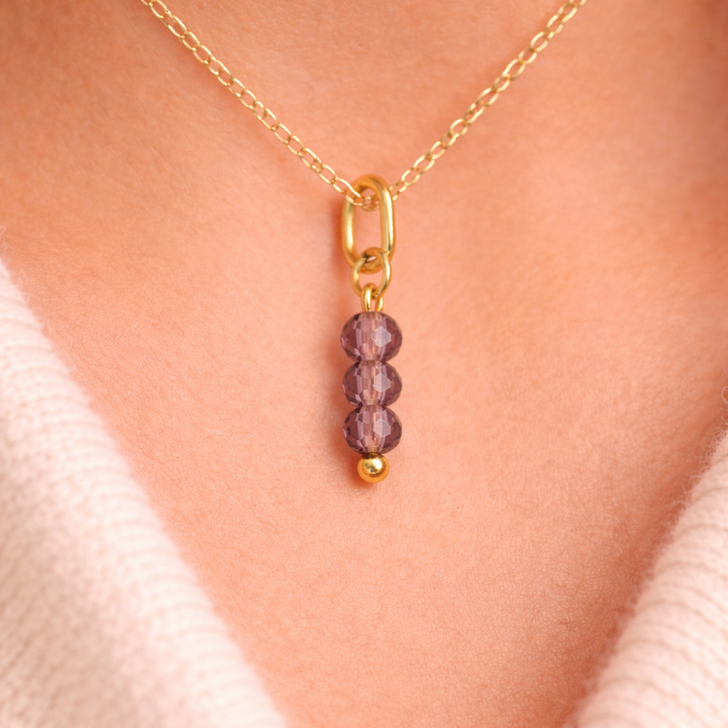 The Three Beads CHARM worn at the neckline, three faceted mauve beads and a gold ball on a gold paperclip chain over a cream sweater.