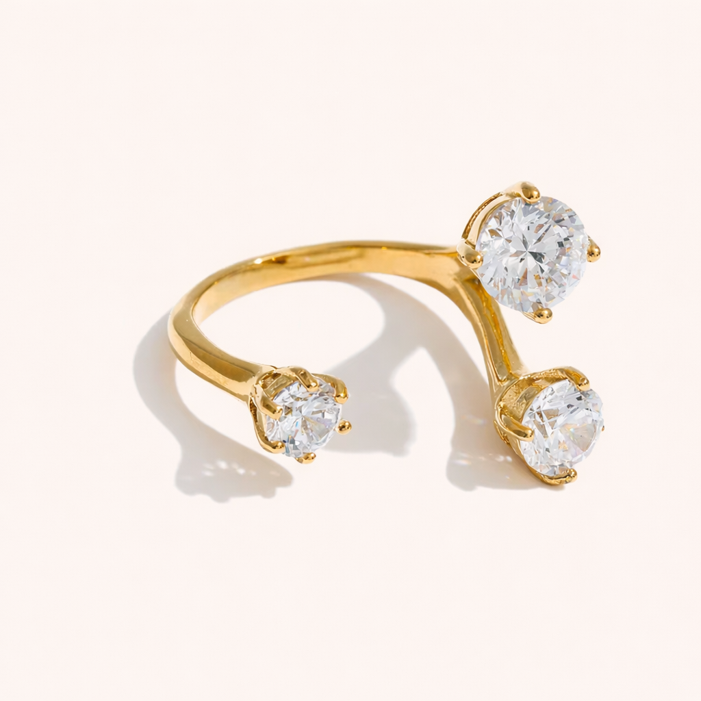 Open gold ring set with three round clear stones at different heights, shown at an angle on a cream backdrop — Trinity RING.