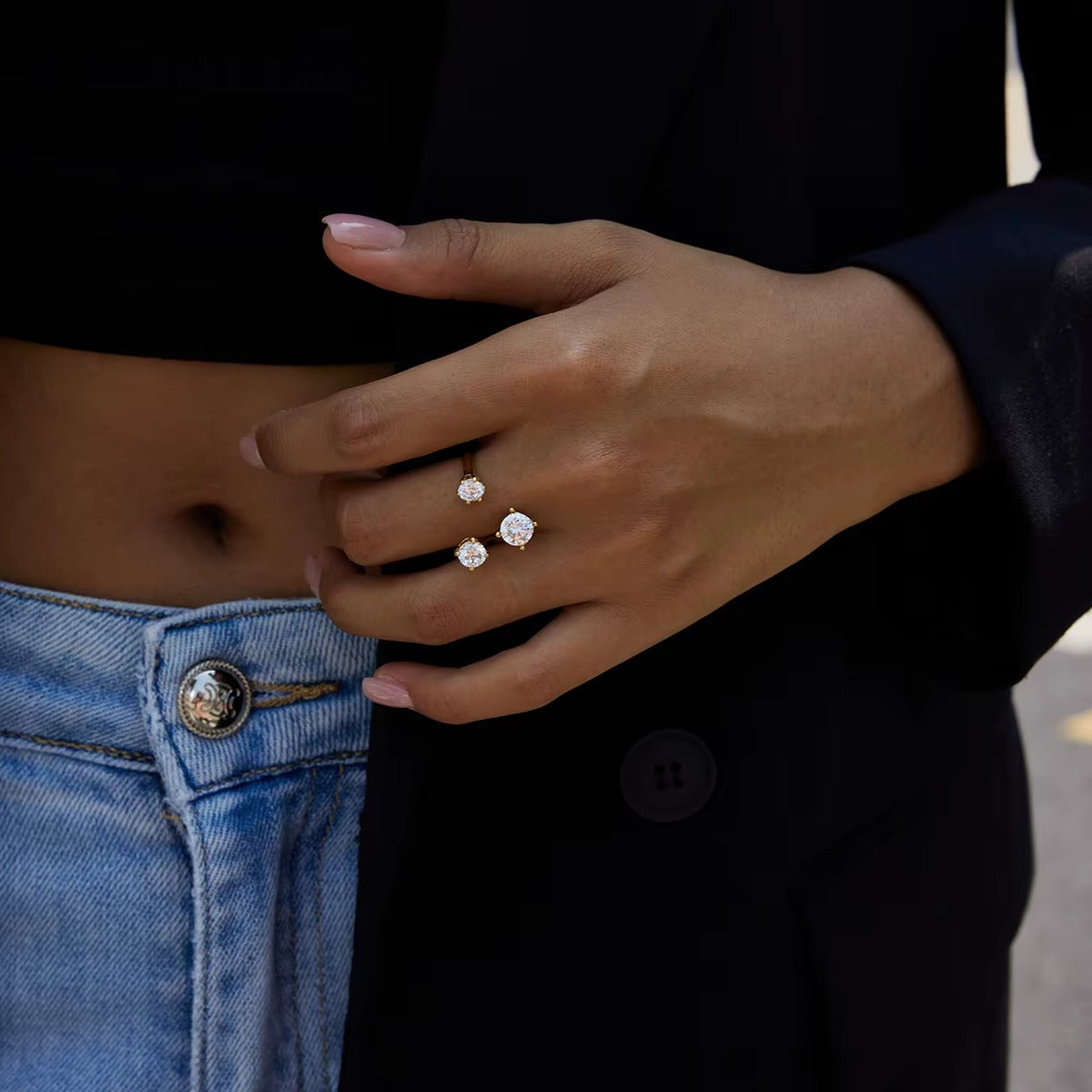 Close-up of a hand wearing the open gold Trinity RING with its three clear stones, resting against a denim waistband.
