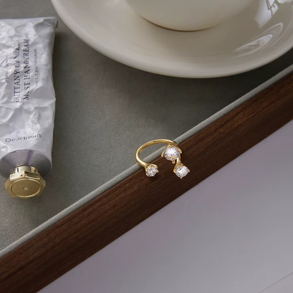 Open gold Trinity RING with three clear stones styled on a wooden tray edge beside a hand cream tube and teacup.