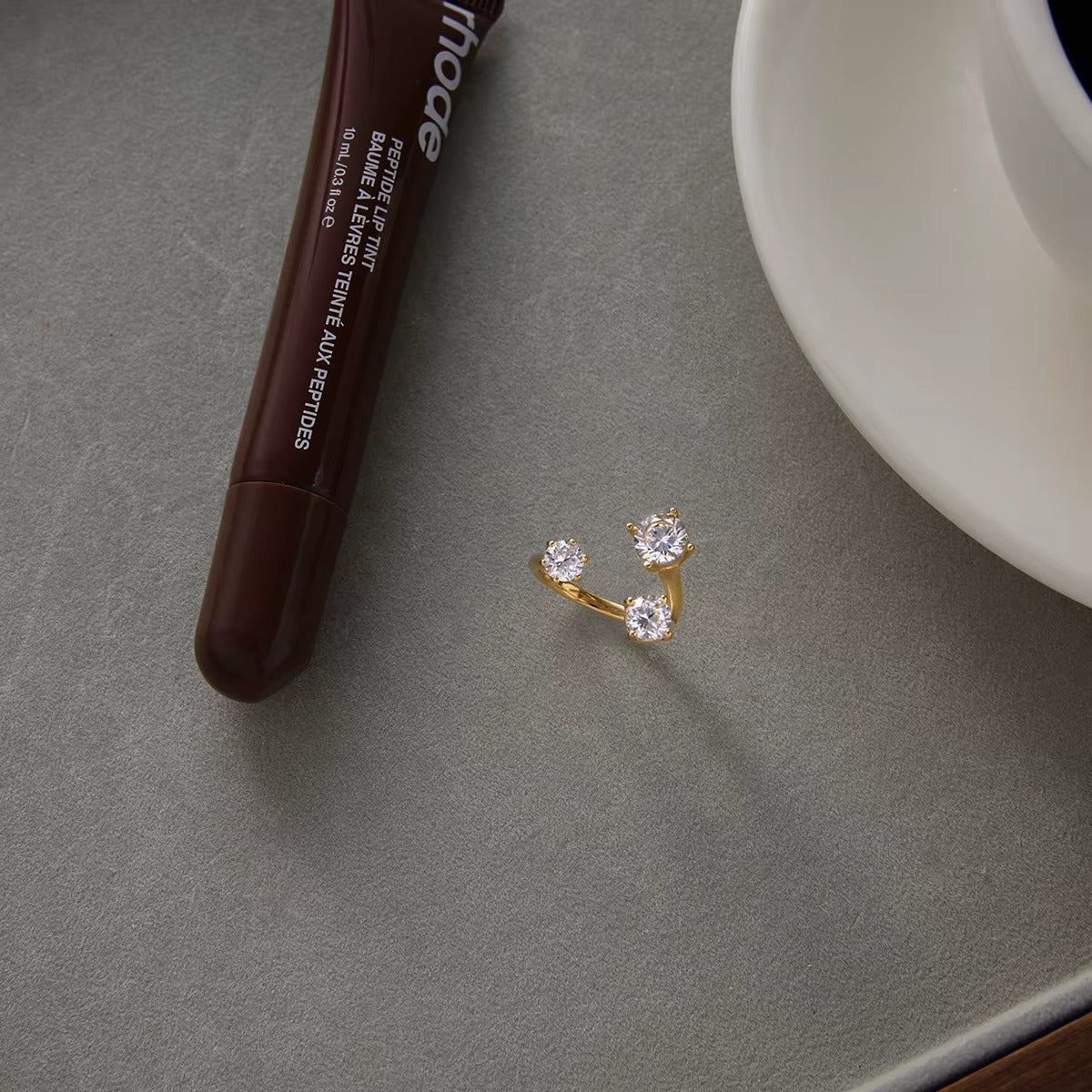 Open gold Trinity RING with three clear stones styled on a grey surface beside a lip tint tube and teacup.