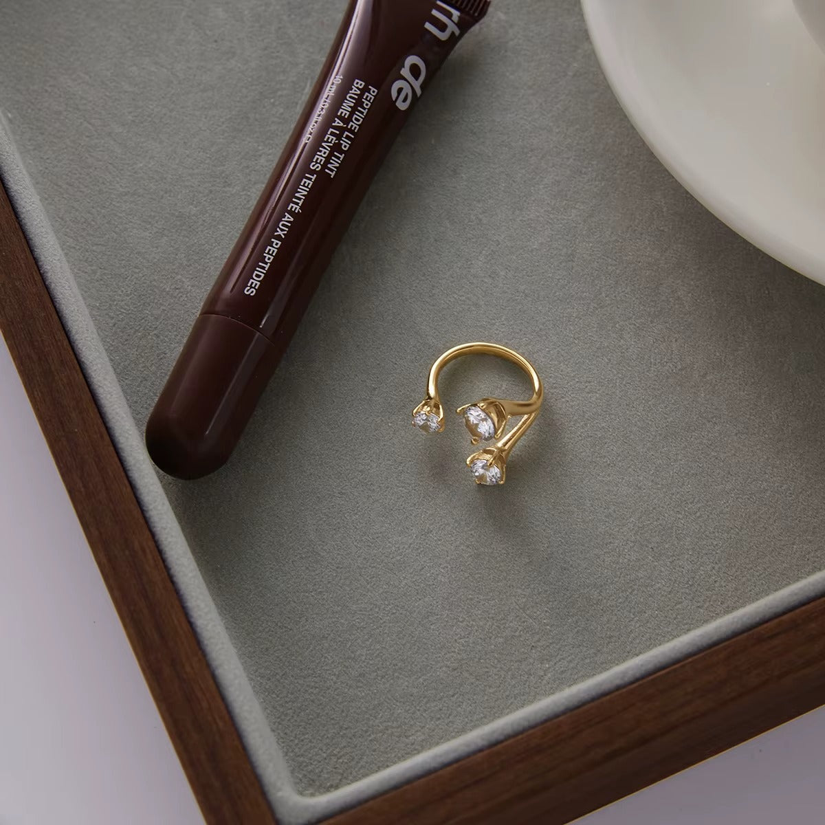 Open gold Trinity RING with three clear stones displayed in a velvet-lined jewelry tray beside a lip tint tube.