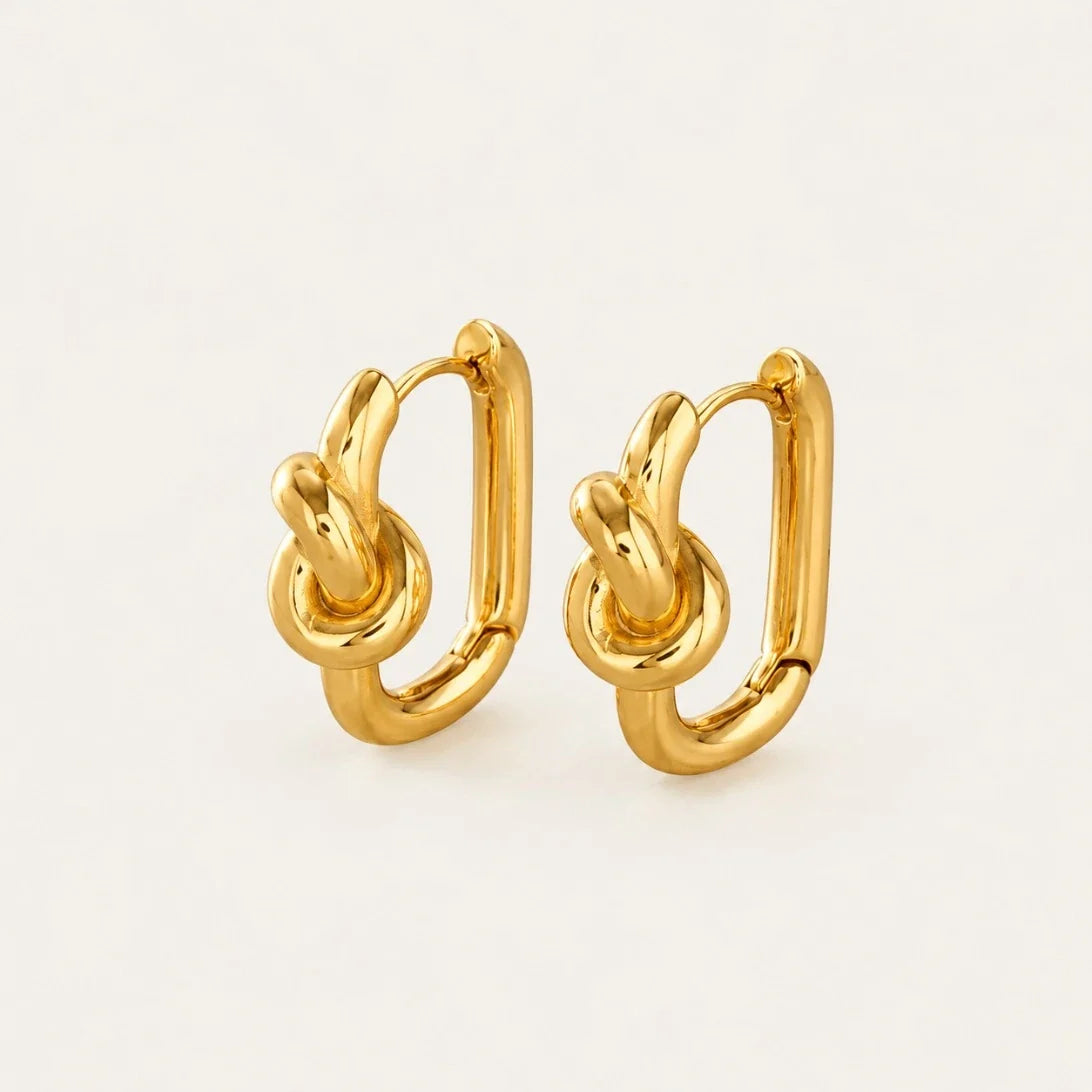 Pair of gold Twine EARRINGS, oval hoops with a twisted knot accent, on a cream background — Folie à Trois
