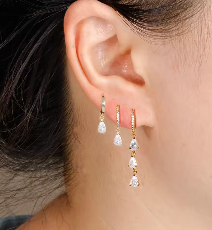 Waterfall Earrings worn on the ear, the cascading pear-crystal drops stacked beside two shorter huggie hoops.