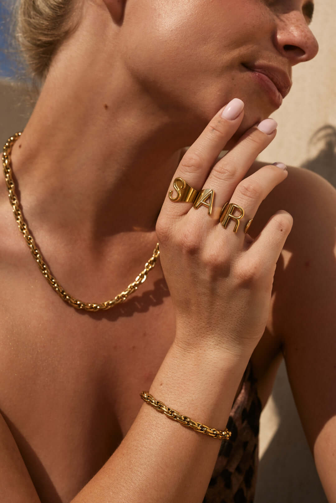 Close-up of a hand spelling out a name with stacked gold letter rings, worn near the chin in bright sunlight.