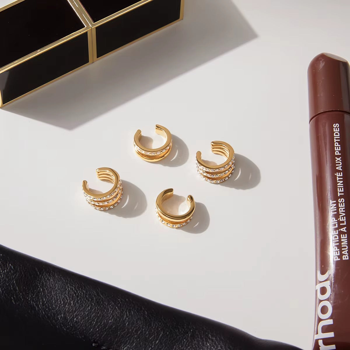 Four Wildcard Ear Cuffs arranged on a white marble surface beside a lip tint tube, each showing the coiled pave crystal spiral design.