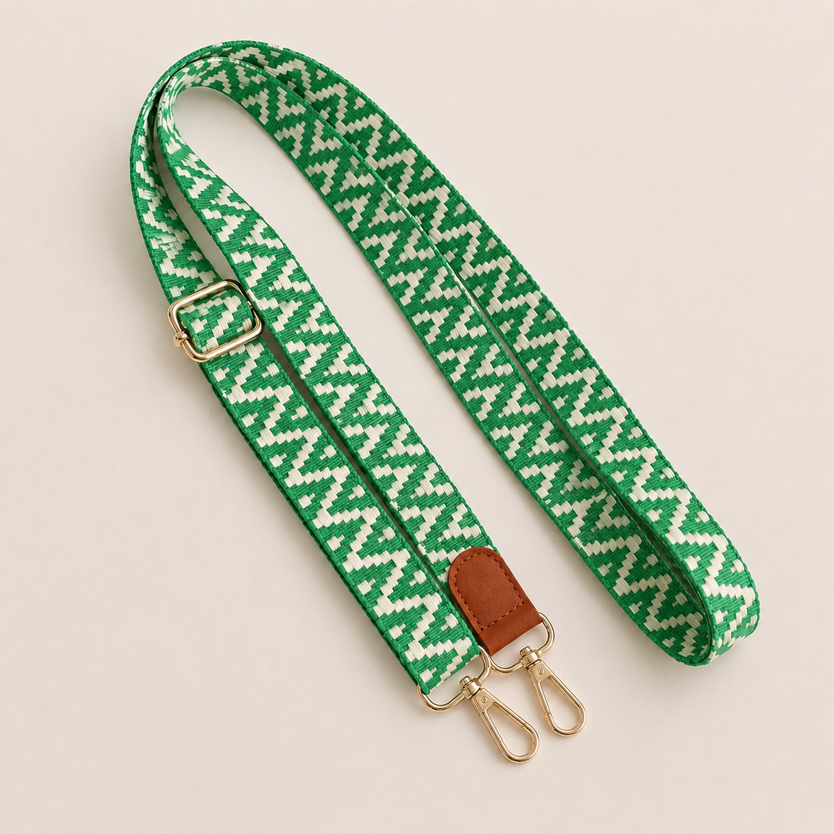 Folie à Trois Wynter long wholesale phone strap in a green and white zigzag weave with a tan leather tab and gold clasps, coiled on a cream background.